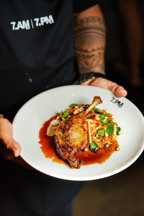 A plate of roasted duck leg with sauce held by a server at 7AM Bakers and 7PM Diners