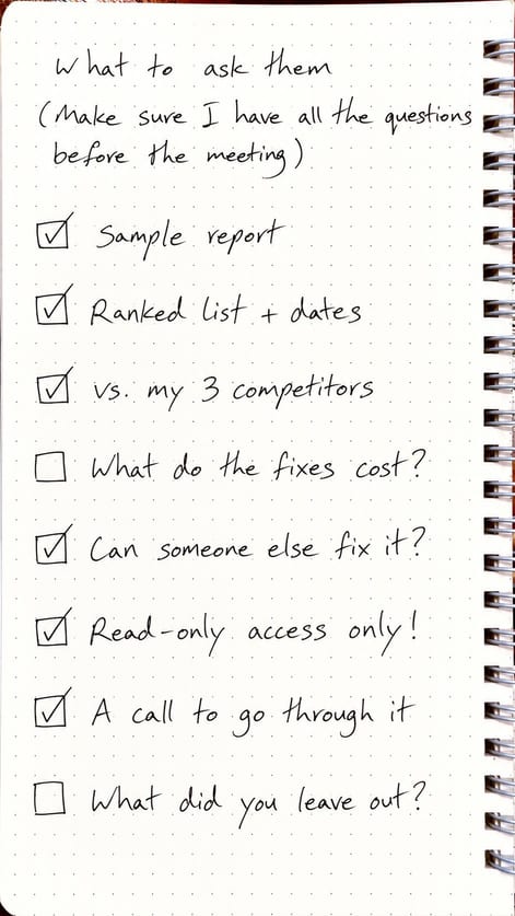 A handwritten checklist in a dot-grid spiral notebook headed "What to ask them (make sure I have all the questions before the meeting)": sample report, ranked list and dates, vs. my 3 competitors, what the fixes cost, can someone else fix it, read-only access, a call to go through it, and what was left out, with six boxes ticked and two still empty